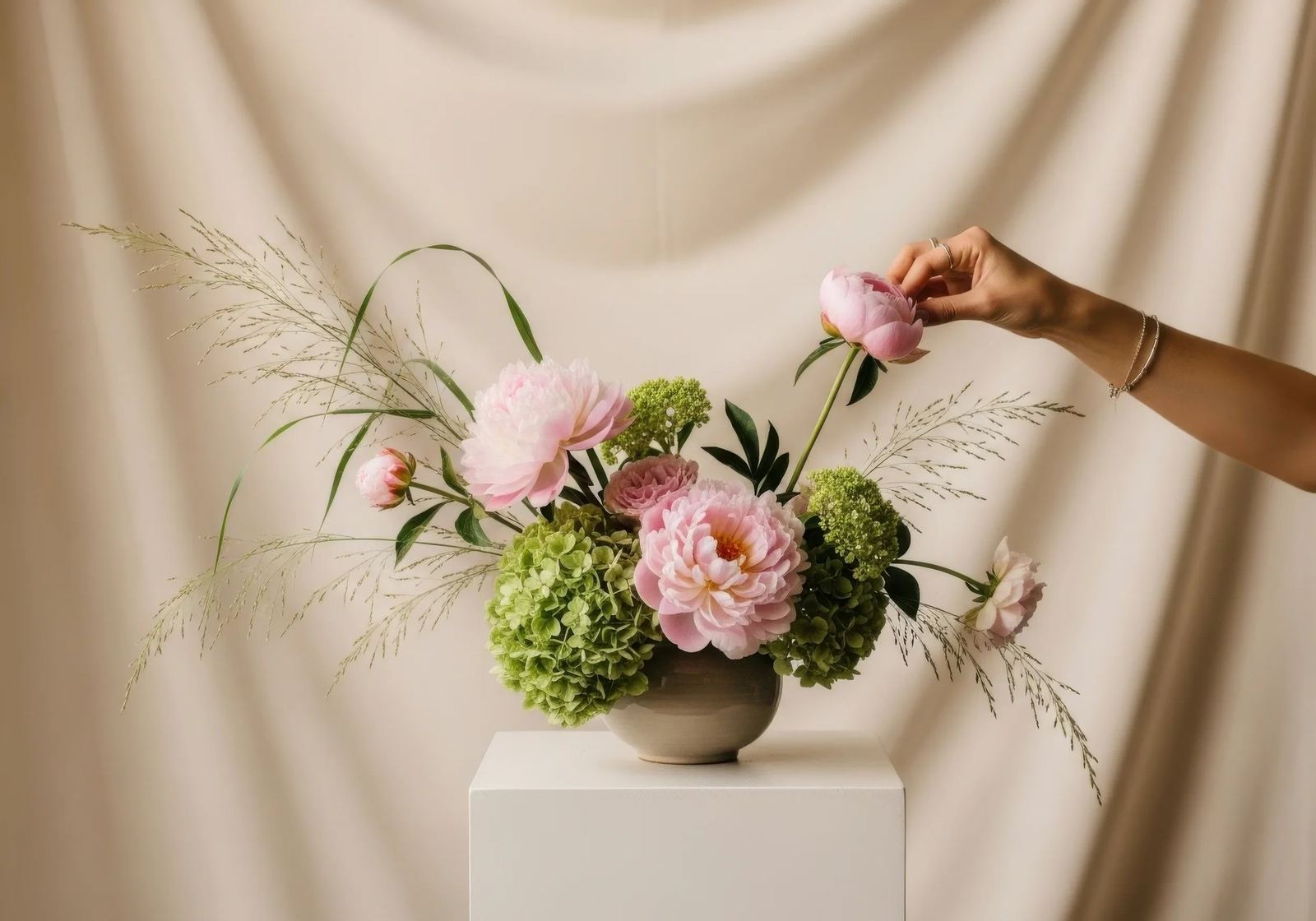 A hand adjusts a pink peony in an arrangement of peonies, green hydrangea and grasses in a stone bowl on a white block, against a draped cream backdrop.