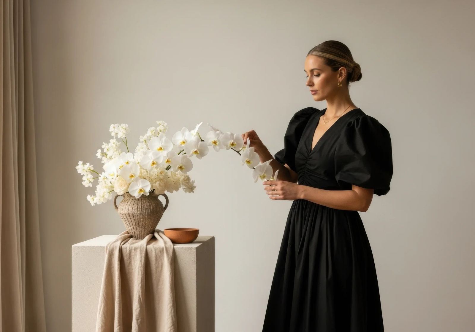 A woman in a black puff-sleeved dress arranges white orchids in a ribbed clay vase on a pale stone pedestal.