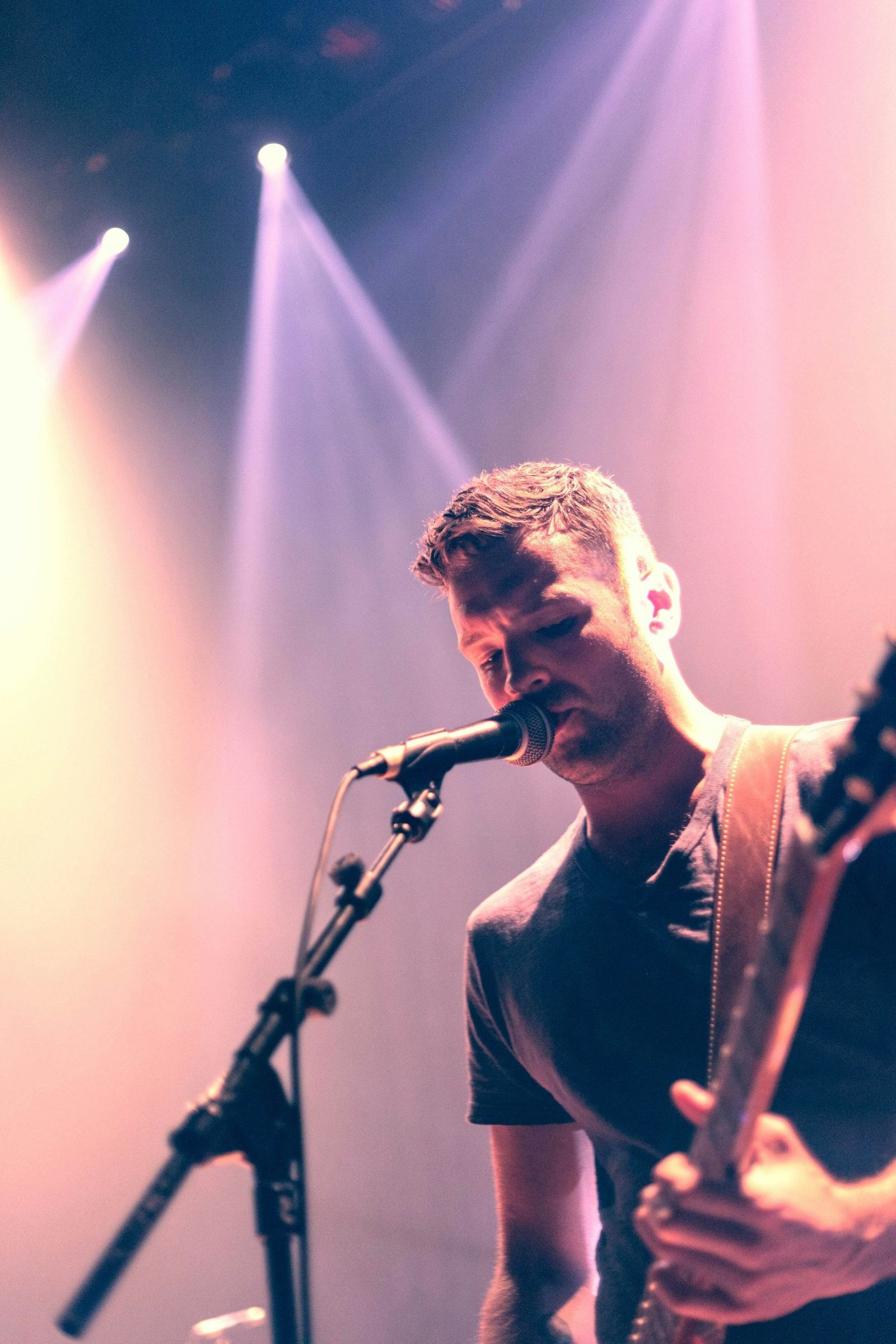 A man with short hair sings into a microphone while holding a guitar, lit by pink and purple stage lights.