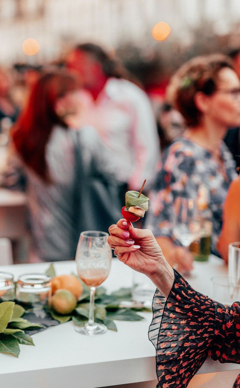 A hand holds a small skewer of tomato, cheese and basil beside a wine glass on a table with leaves and citrus fruit, with guests blurred in the background.