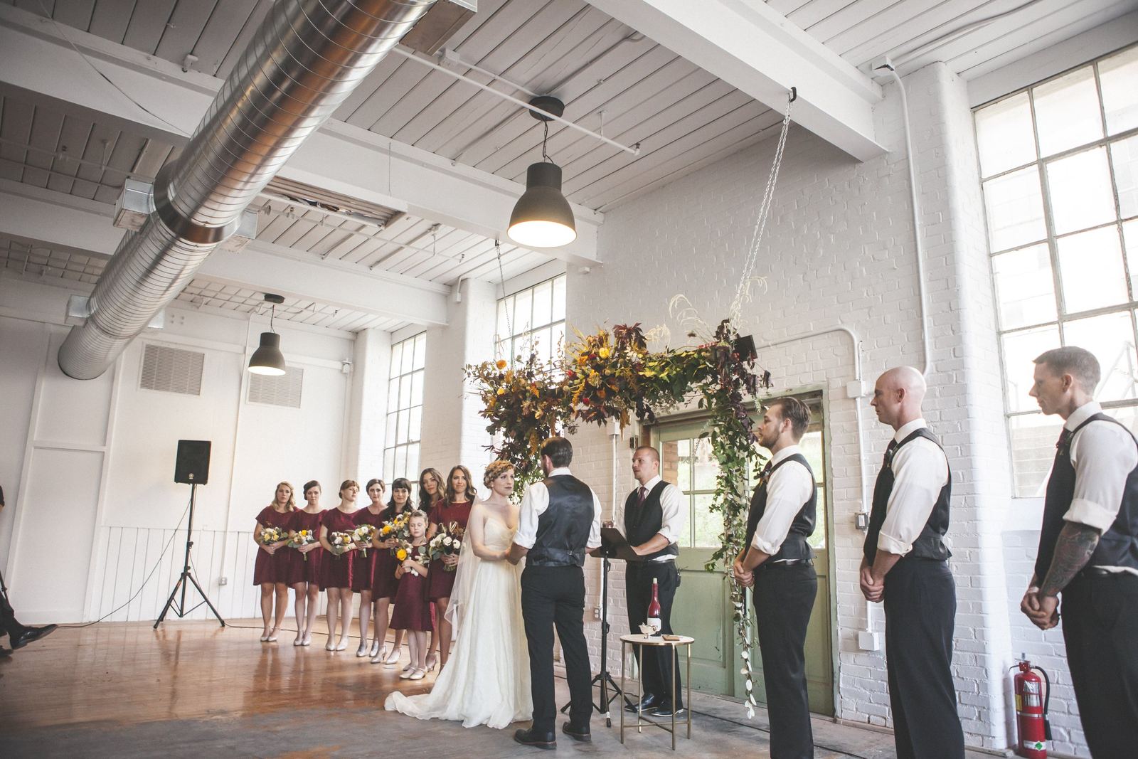 A wedding ceremony inside a bright, industrial-style venue with large windows and white brick walls. The bride and groom face each other under a floral arch, with bridesmaids holding bouquets on the left and groomsmen standing on the right.