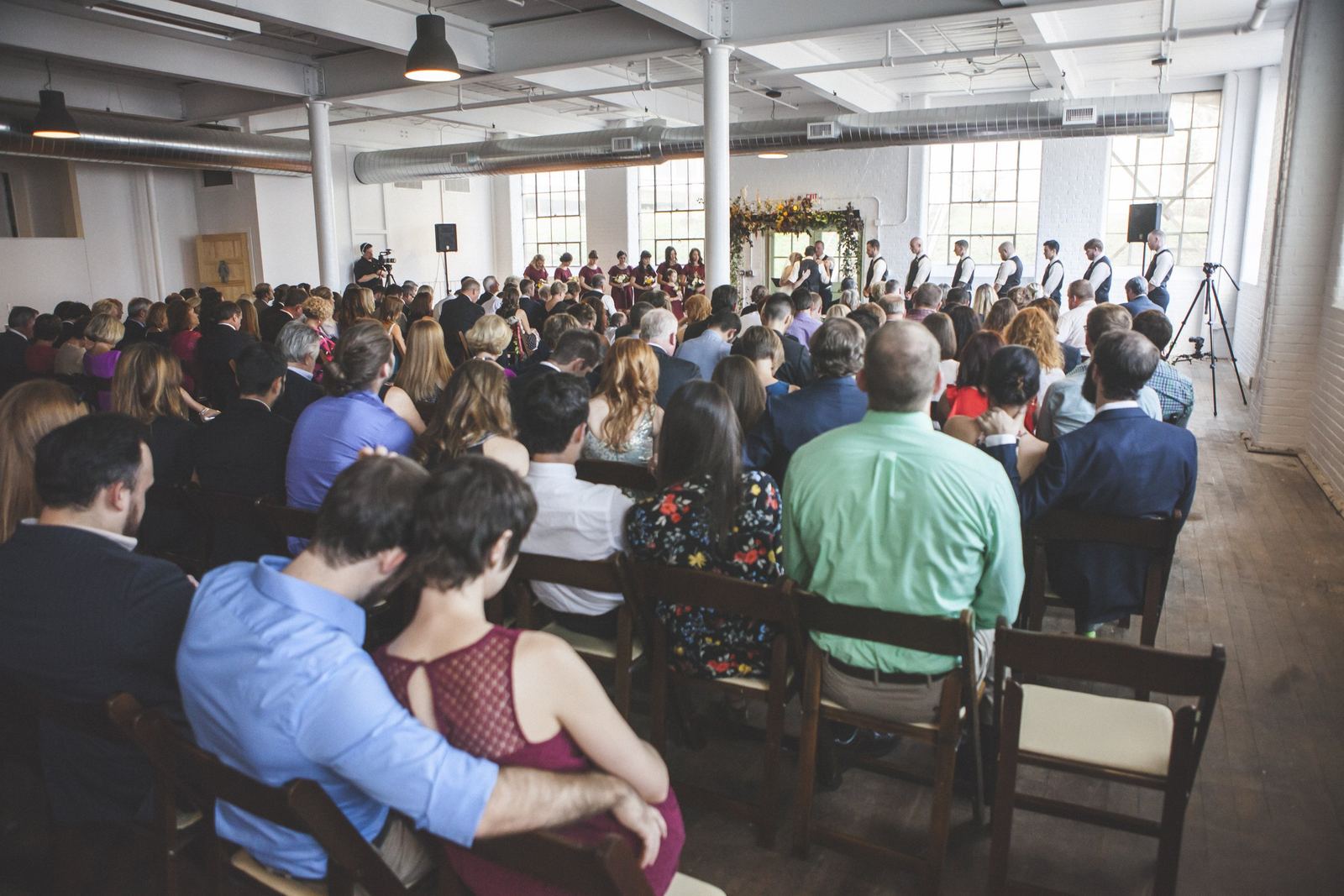 Guests seated in rows of wooden folding chairs face a wedding ceremony at the far end of a bright hall with white brick walls and large windows.